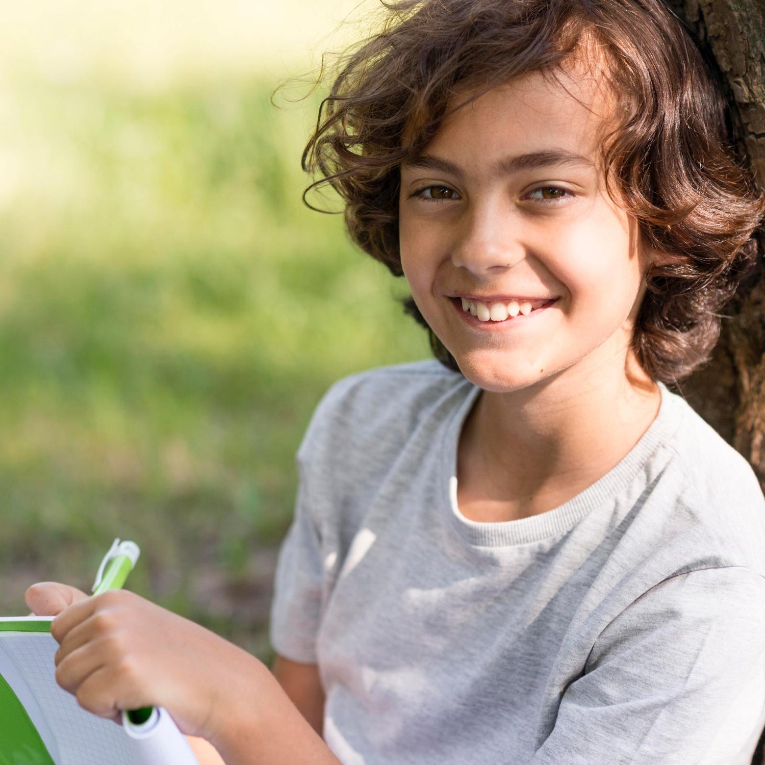 Smiling boy aged 10 leaning against a tree and writing in a journal 