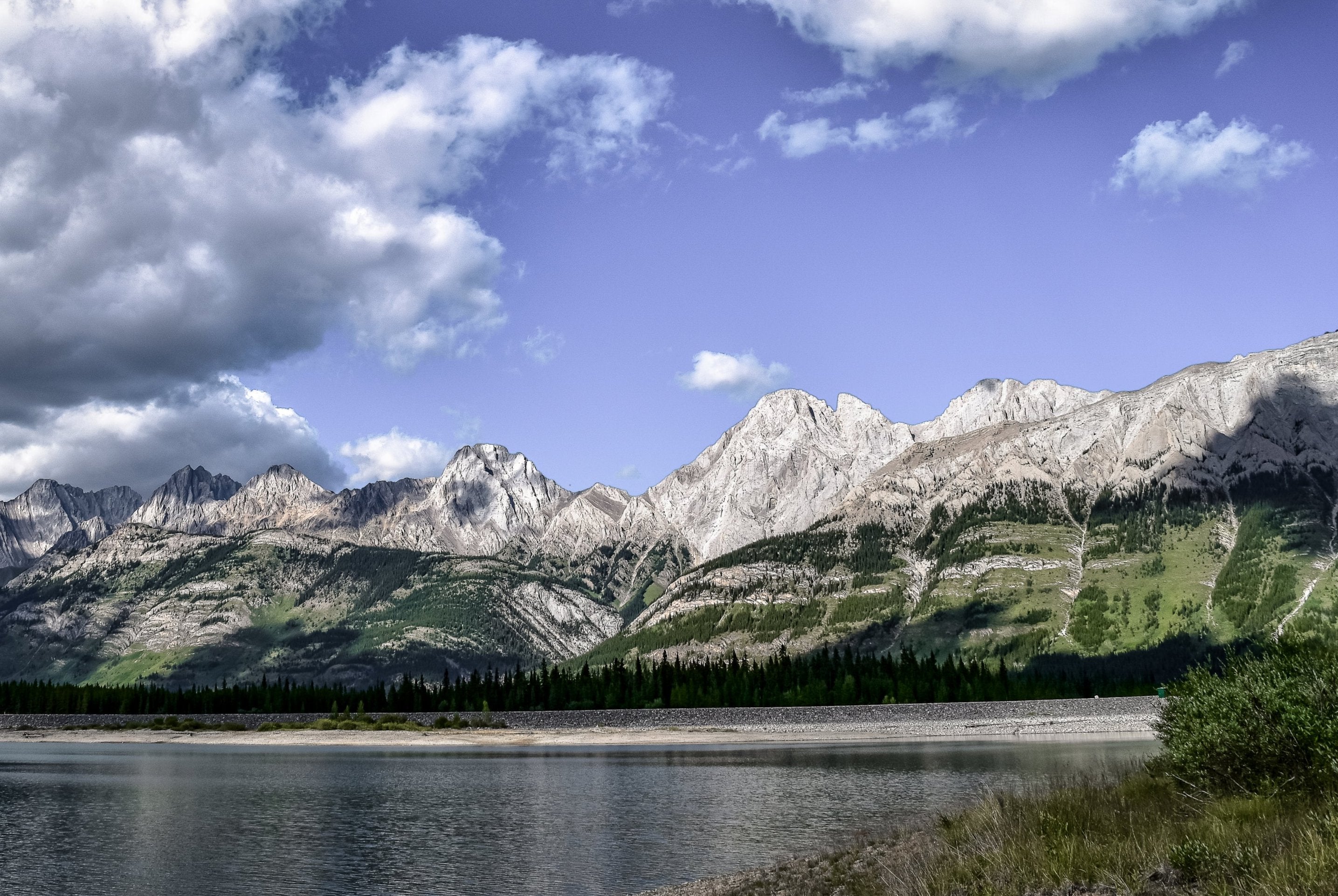 A lake with a mountain range behind with blue skies and white clouds.